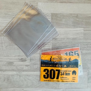 May include: A stack of clear plastic sleeves for protecting and displaying race bibs. The sleeves are on a wood surface. A race bib with the number 307 is visible in the sleeve. The bib is yellow with black text and a red and white graphic. The text on the bib reads "полудиво трасе 34 km".