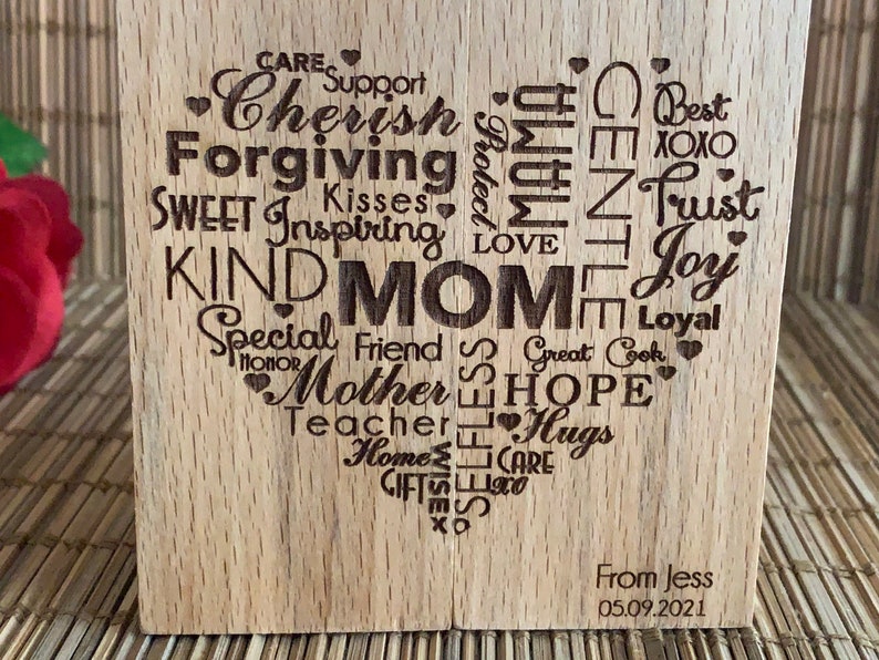 May include: Wooden heart-shaped plaque with the word "MOM" in the center surrounded by words describing a mother's qualities, such as "Cherish", "Forgiving", "Kind", "Special Friend", "Hope", "Love", "Trust", "Joy", and "Hugs". The plaque also includes the text "From Jess 05.09.2021".