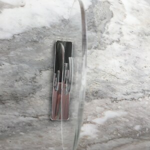 May include: A clear plastic adhesive hook with a chrome finish. The hook is mounted on a white marble surface.