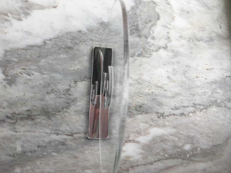 May include: A clear plastic adhesive hook with a chrome finish. The hook is mounted on a white marble surface.