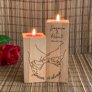 May include: Two wooden candle holders with a hand-drawn design of two hands holding each other. The candle holders are engraved with the text "Jasmine & Robert May 5th 2021 Forever & always".