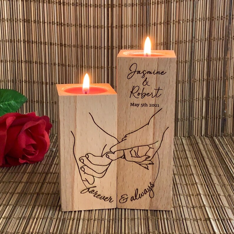 May include: Two wooden candle holders with a hand-drawn design of two hands holding each other. The candle holders are engraved with the text "Jasmine & Robert May 5th 2021 Forever & always".