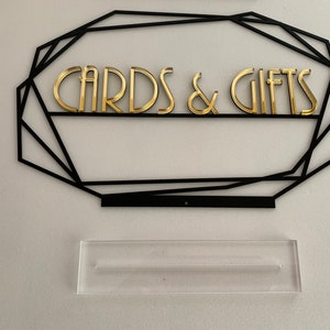 K&ouml;nnte beinhalten: Ein schwarzer geometrischer Rahmen mit goldenen Buchstaben, auf dem "CARDS & GIFTS" steht. Unter dem Rahmen befindet sich ein durchsichtiger Acryl-St&auml;nder.
