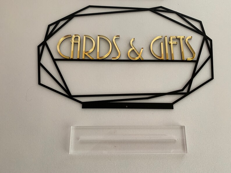 K&ouml;nnte beinhalten: Ein schwarzer geometrischer Rahmen mit goldenen Buchstaben, auf dem "CARDS & GIFTS" steht. Unter dem Rahmen befindet sich ein durchsichtiger Acryl-St&auml;nder.