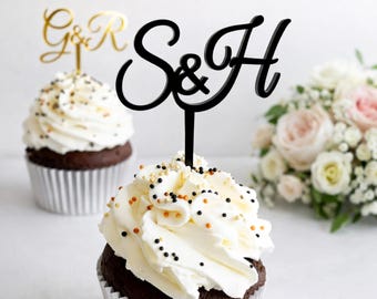 May include: Two cupcake toppers featuring cursive lettering. One topper is gold and displays the letters G & R. The second topper is black and displays the letters S & H. Each topper has a thin stem for insertion into a cupcake with white frosting.