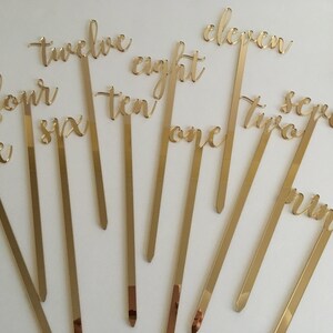 May include: Gold acrylic table numbers on sticks, with numbers one through twelve. The numbers are written in a cursive script.