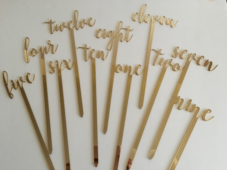 May include: Gold acrylic table numbers on sticks, with numbers one through twelve. The numbers are written in a cursive script.