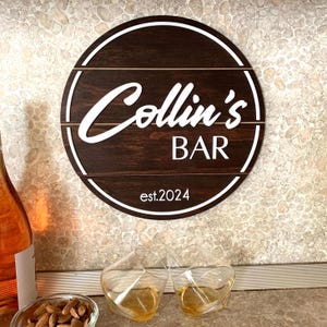 Peut inclure: Une enseigne ronde en bois avec le texte "Collin's Bar est. 2024" en lettres blanches. L'enseigne a une finition en bois brun foncé et une bordure blanche.