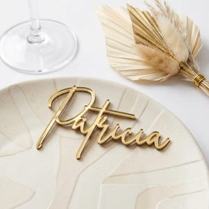 Könnte beinhalten: Ein goldfarbener Namensschild mit dem Namen "Patricia" in eleganter Schreibschrift liegt auf einem cremefarbenen Teller. Ein Weinglas und ein Trockenblumenarrangement befinden sich im Hintergrund. Ideal für Hochzeiten oder Veranstaltungen.