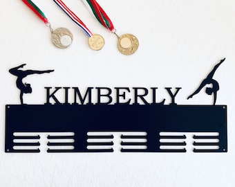 May include: Black metal medal display with the name "KIMBERLY" and a silhouette of a gymnast on a balance beam. The display has 12 slots for medals and is designed to hang on a wall.