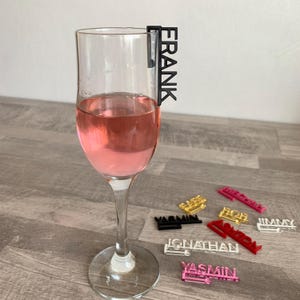 A set of colorful acrylic drink markers with names printed on them. The names include Frank, Yasmin, Jonathan, Abigail, Rich, Jimmy, and Basil. The markers are designed to be placed on the rim of a glass to identify drinks.
