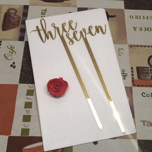 May include: Gold 'three seven' cake topper with two gold sticks. The topper is on a white surface with a red rose.