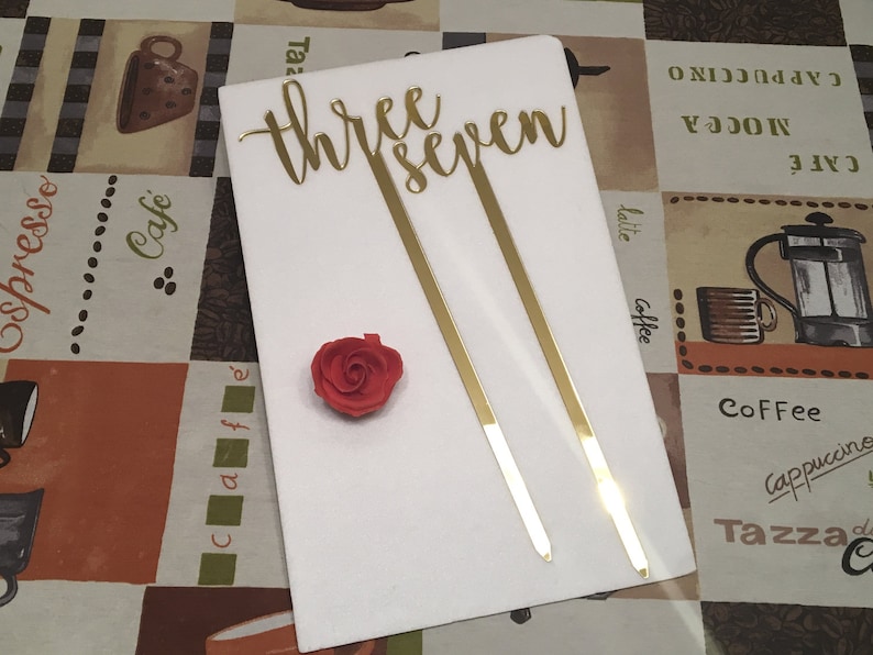 May include: Gold 'three seven' cake topper with two gold sticks. The topper is on a white surface with a red rose.
