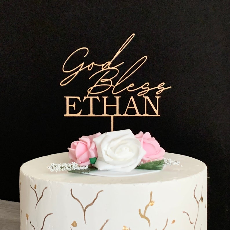以下が含まれることがあります： 金箔のアクセントが施された白いケーキの上に、「God Bless Ethan」と金色の文字で書かれた木製ケーキトッパーが置かれています。トッパーはピンクと白のバラで囲まれています。