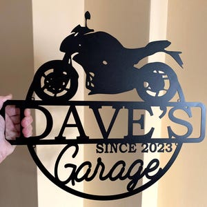 K&ouml;nnte beinhalten: Schwarzes Metallschild mit einer Motorradsilhouette, dem Text "Dave's Garage Since 2023" und einem runden Rand.