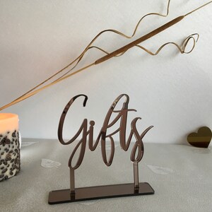 May include: A rose gold mirrored acrylic sign with the word "Gifts" in a cursive font. The sign is on a stand with a mirrored base.