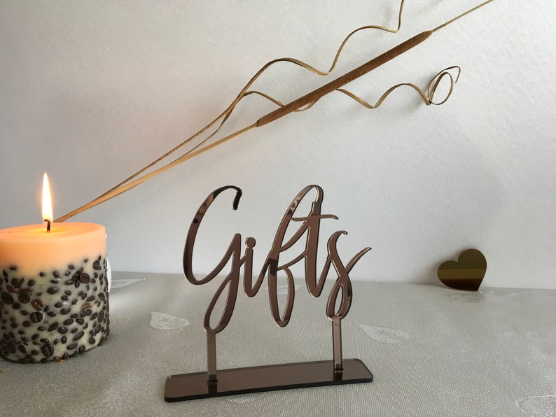 May include: A rose gold mirrored acrylic sign with the word "Gifts" in a cursive font. The sign is on a stand with a mirrored base.