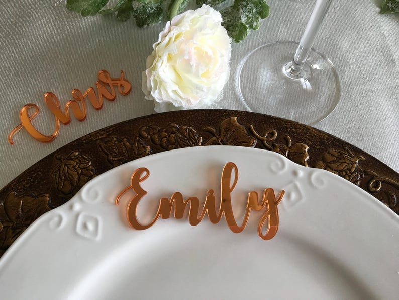 Peut inclure: Deux marque-places personnalis&eacute;s en acrylique orange r&eacute;fl&eacute;chissant, portant les pr&eacute;noms Emily et Chris en &eacute;criture cursive, pos&eacute;s sur une assiette blanche avec un dessous d'assiette d&eacute;coratif en bronze.