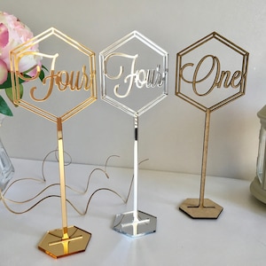 May include: Three gold, silver, and wood table number holders with geometric hexagonal frames. The numbers "Four", "Four", and "One" are written in a cursive font on the holders.