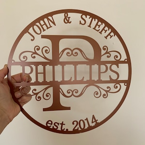 May include: A copper-colored metal family name sign. The sign is round with the names "JOHN & STEFF" at the top and "PHILLIPS" in the center. The letter "P" is the central design element, with decorative flourishes. The year "est. 2014" is at the bottom.