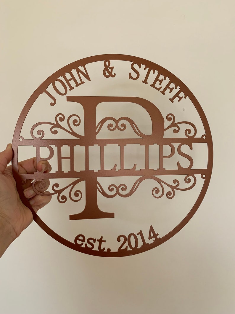 May include: A copper-colored metal family name sign. The sign is round with the names "JOHN & STEFF" at the top and "PHILLIPS" in the center. The letter "P" is the central design element, with decorative flourishes. The year "est. 2014" is at the bottom.