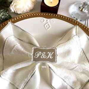 May include: A white napkin with a silver trim is folded into a bow shape and held together with a silver napkin ring. The ring has a rectangular plaque with the initials "P&K" in a white font.