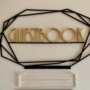 K&ouml;nnte beinhalten: Ein schwarzer geometrischer Rahmen mit dem Wort "GUESTBOOK" in goldenen Buchstaben. Unter dem Rahmen befindet sich ein durchsichtiger Acryl-St&auml;nder.