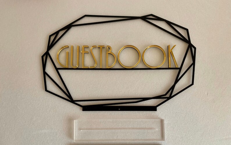 K&ouml;nnte beinhalten: Ein schwarzer geometrischer Rahmen mit dem Wort "GUESTBOOK" in goldenen Buchstaben. Unter dem Rahmen befindet sich ein durchsichtiger Acryl-St&auml;nder.