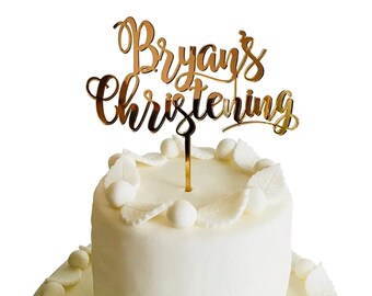 May include: Gold glitter cake topper with the text "Bryan's Christening" on a white cake.