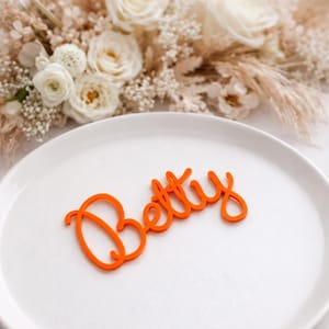 Peut inclure: Une &eacute;tiquette nominative en acrylique orange vif portant le pr&eacute;nom Betty &eacute;crit dans une police cursive. Le nom d&eacute;coup&eacute; est plac&eacute; au centre d'une assiette blanche lisse et unie, cr&eacute;ant un contraste net entre l'orange et le blanc.