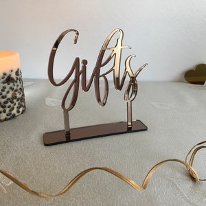 May include: Rose gold mirrored acrylic sign with the word "Gifts" in a cursive font. The sign is on a table with a candle and a gold heart.