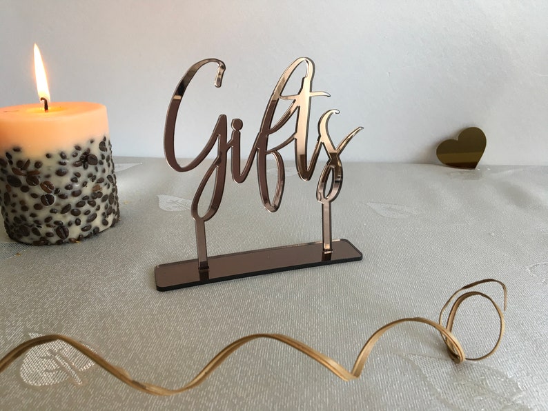 May include: Rose gold mirrored acrylic sign with the word "Gifts" in a cursive font. The sign is on a table with a candle and a gold heart.