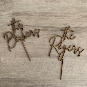 May include: Two wooden cake toppers with the words "The Beckers" and "The Rogers" in a cursive font. Each topper has a stake for insertion into a cake. The toppers are made of light brown wood.