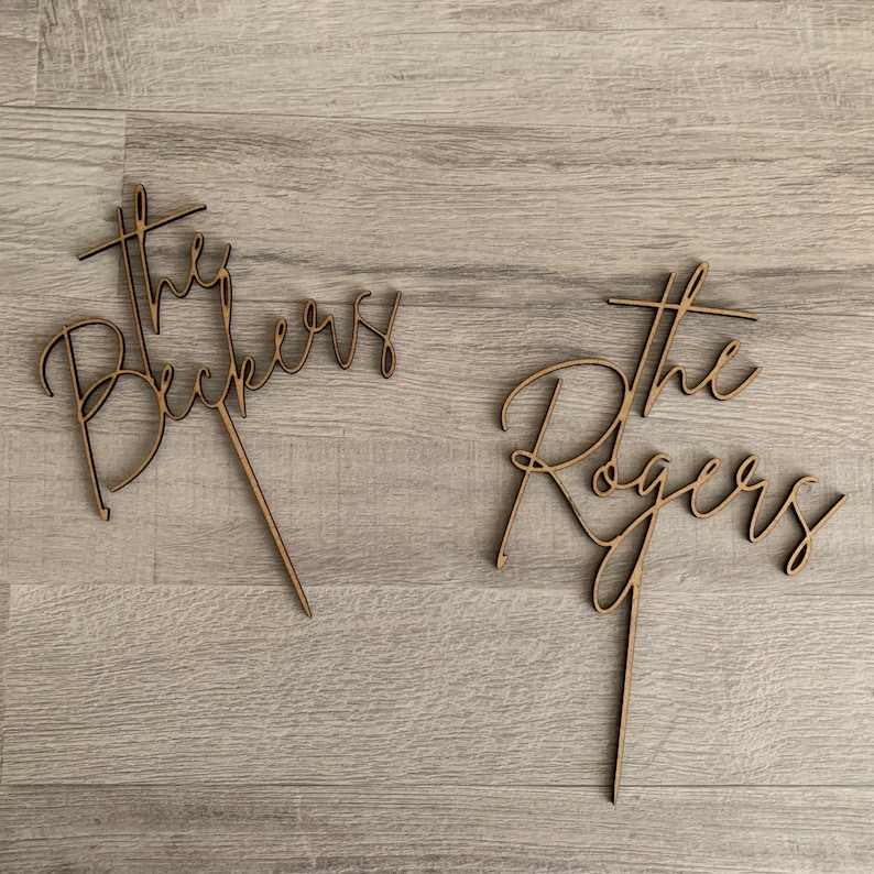 May include: Two wooden cake toppers with the words "The Beckers" and "The Rogers" in a cursive font. Each topper has a stake for insertion into a cake. The toppers are made of light brown wood.