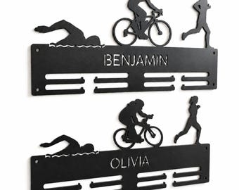 Op de afbeelding: Twee zwarte metalen medaillehangers voor aan de muur met silhouetten van een zwemmer, fietser en hardloper. De bovenste hanger toont de naam BENJAMIN en de onderste de naam OLIVIA, beide voorzien van horizontale sleuven voor medailles.