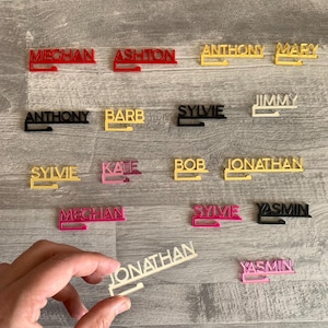 A collection of personalized name hooks in various colors, including black, red, pink, yellow, and cream. The hooks are made of plastic and have a simple design. The names on the hooks include: Meghan, Ashton, Anthony, Mary, Jimmy, Barb, Sylvie, Kate, Bob, Jonathan, Yasmin.