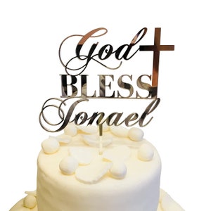 Pode incluir: Topper para bolo de acrílico espelhado dourado com o texto "God Bless Jonael" e uma cruz. O topper está em um bolo branco com cobertura branca e decorações brancas.
