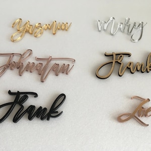 May include: Assortment of acrylic table place cards with names written in cursive. The names include "Yazmin", "Mary", "Jonathan", "Frank", and "Love". The cards are in various colours, including gold, silver, black, and rose gold.
