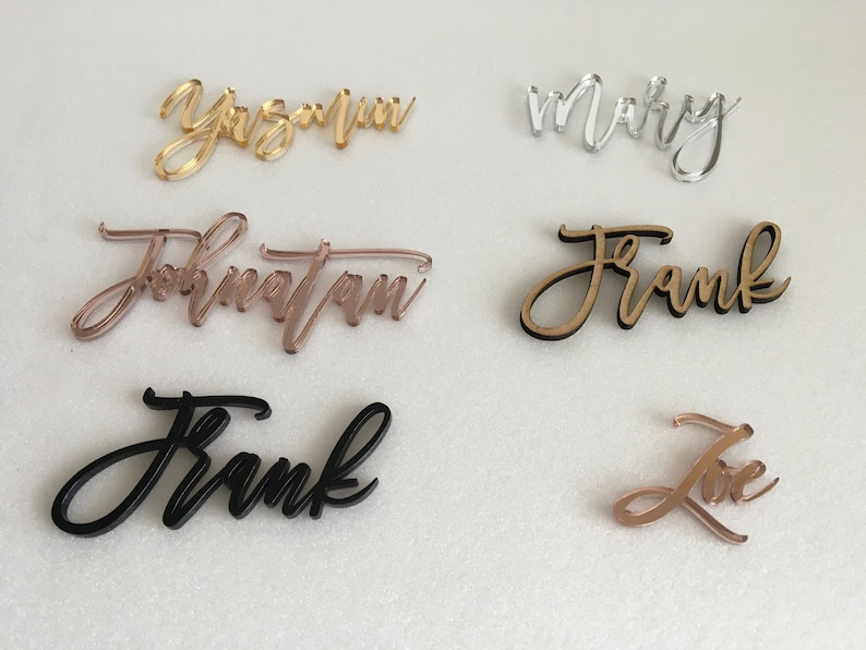 May include: Assortment of acrylic table place cards with names written in cursive. The names include "Yazmin", "Mary", "Jonathan", "Frank", and "Love". The cards are in various colours, including gold, silver, black, and rose gold.