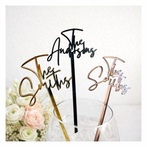 May include: Three cake toppers with the words "The Smiths" and "The Andersons" in elegant script. One is gold, one is black, and one is rose gold. They are displayed in a clear glass with flowers in the background.