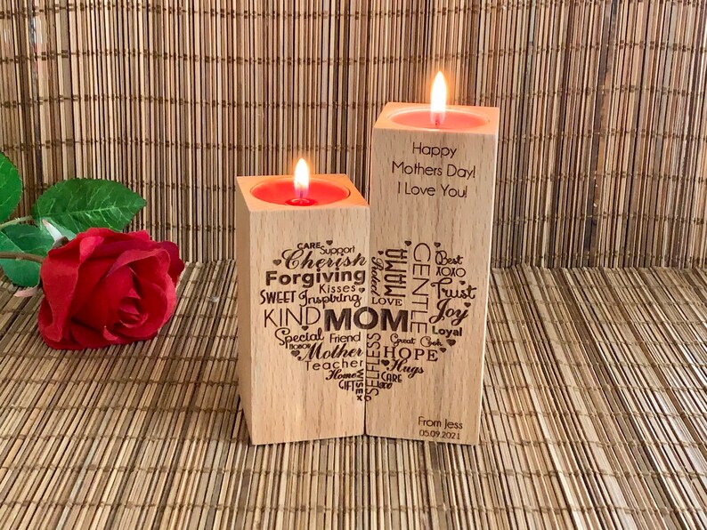 May include: Two wooden candle holders with a heart-shaped design. The left holder has words like "Cherish", "Forgiving", "Kind Mom", and "Special Friend" engraved on it. The right holder has the text "Happy Mothers Day I Love You" engraved on it. The candle holders are on a woven bamboo surface.