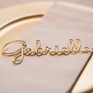 May include: Gold acrylic place card with the name "Gabriella" written in cursive.