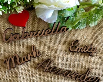 May include: Wooden place cards with the names Gabriella, Mark, Emily, and Alexander written in cursive. The place cards are on a brown burlap surface.