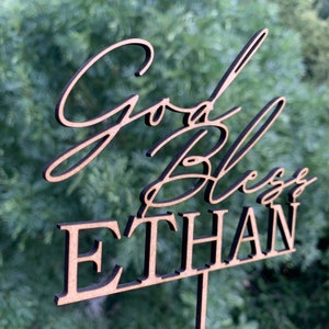 以下が含まれることがあります： 黒い筆記体で「God Bless Ethan」と書かれた木製ケーキトッパー