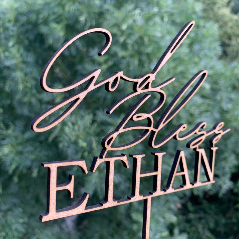 以下が含まれることがあります： 黒い筆記体で「God Bless Ethan」と書かれた木製ケーキトッパー