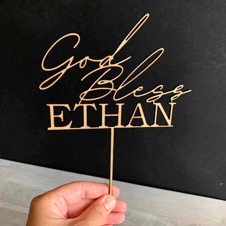 以下が含まれることがあります： 「God Bless Ethan」というテキストが書かれた木製のケーキトッパー。筆記体で書かれており、トッパーは茶色で、下部に棒が取り付けられています。