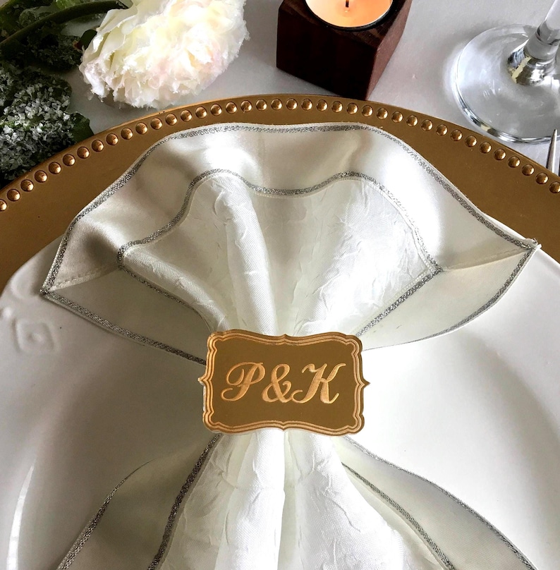 May include: Gold napkin ring with a rectangular design and the initials "P&K" in a cursive font. The ring is placed on a white napkin with a silver trim.