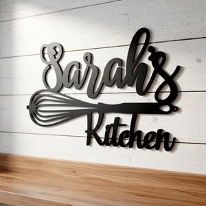 May include: Black metal kitchen sign with the name "Sarah's" above the word "Kitchen". The design includes a whisk incorporated into the lettering, mounted on a white wooden plank wall.