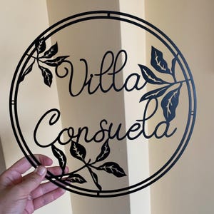 May include: Black metal circular sign with the words "Villa Consuela" in a decorative script font. The sign is decorated with cut-out leaf designs.