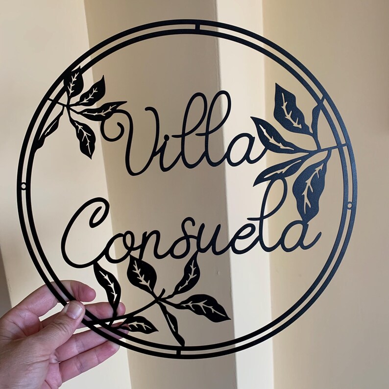May include: Black metal circular sign with the words "Villa Consuela" in a decorative script font. The sign is decorated with cut-out leaf designs.